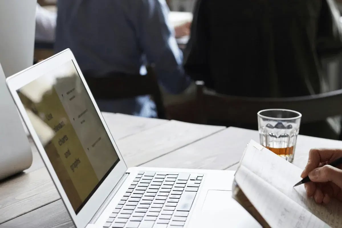 A laptop on a wooden table displays notes, while a person writes in a notebook, with a glass of drink nearby and others in the background.