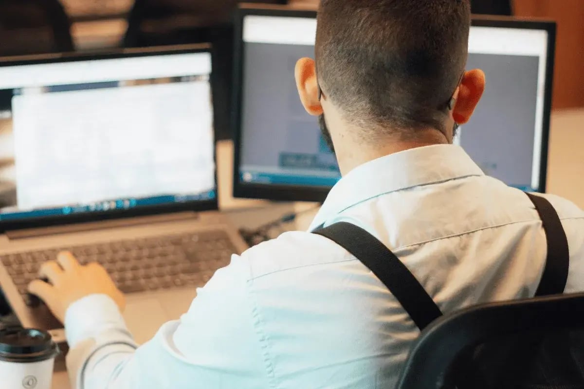 Professional working on laptop with dual monitors while reviewing data in office.