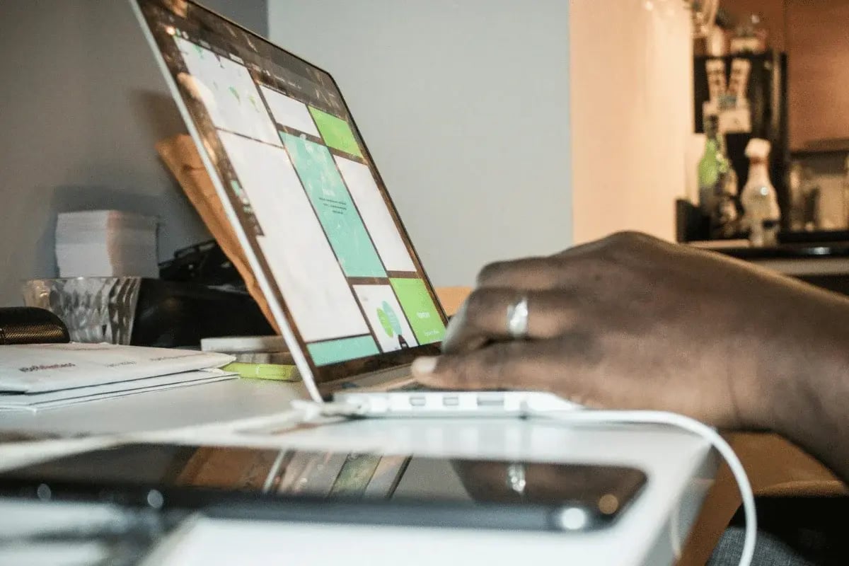 A close-up of a person's hand typing on a laptop at a desk, with a phone and papers scattered around.