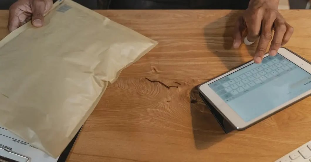 Person holding padded mailer while reviewing amazon PPC optimization metrics on tablet at wooden desk.