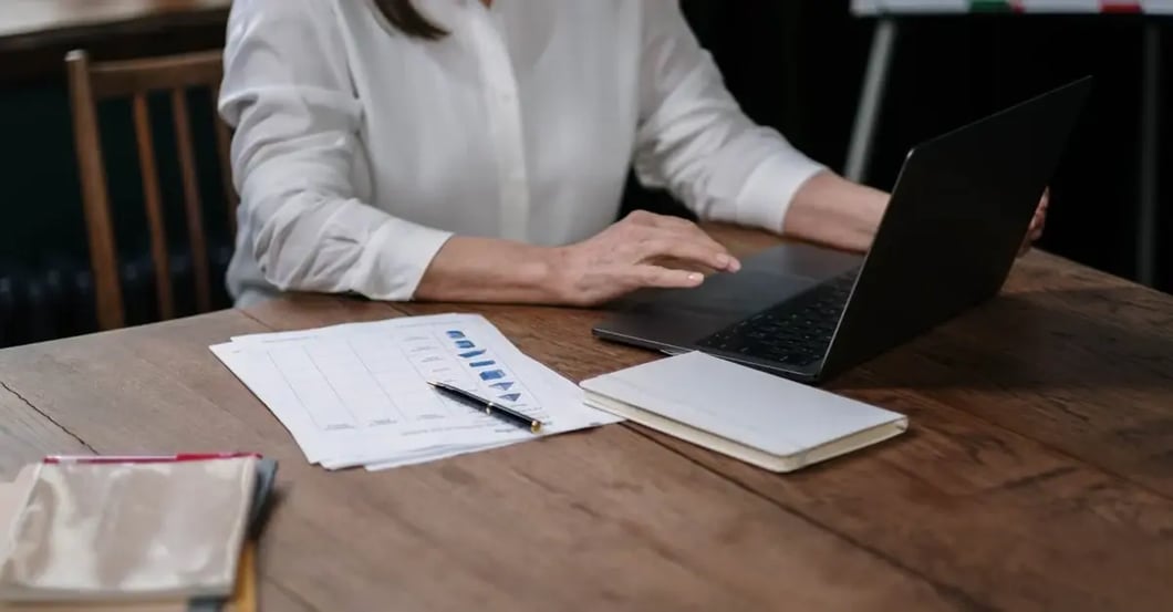 Professional woman using laptop at desk with spreadsheet documents and notepad for amazon flat files data entry.