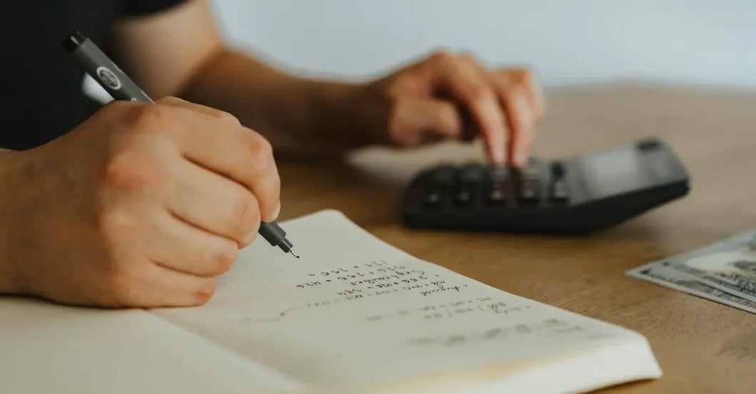 Person calculating amazon PPC management cost figures on notepad at desk with calculator and keyboard nearby.