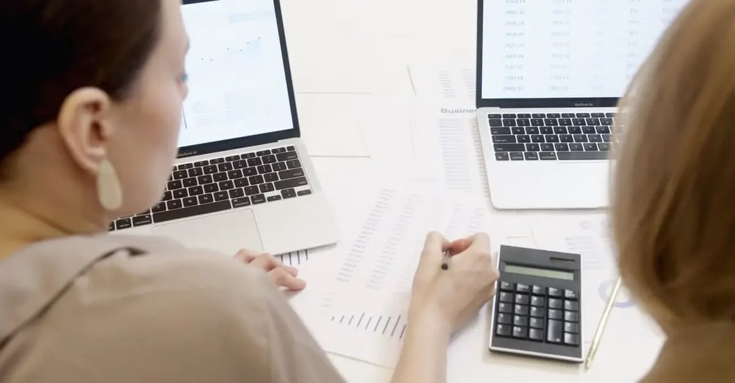 Two marketers analyzing search term reports with laptops and calculator to identify Amazon negative keywords strategy.