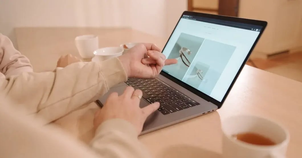 Hands in beige sleeves browse product photos on a laptop at a cozy table, with teacups nearby, creating a calm, focused shopping atmosphere.