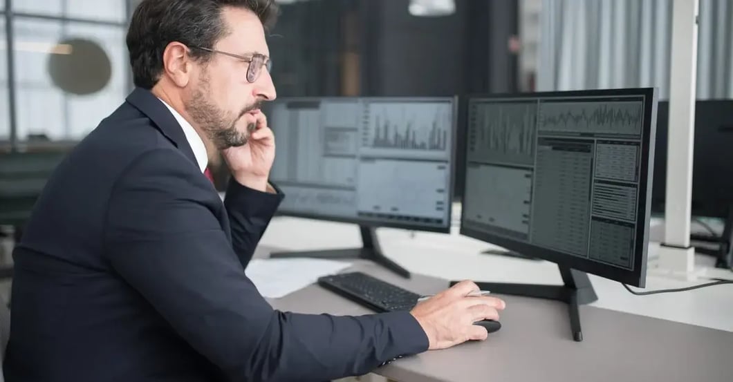 Analyst reviewing Amazon PPC management cost data on dual monitors in office setting.