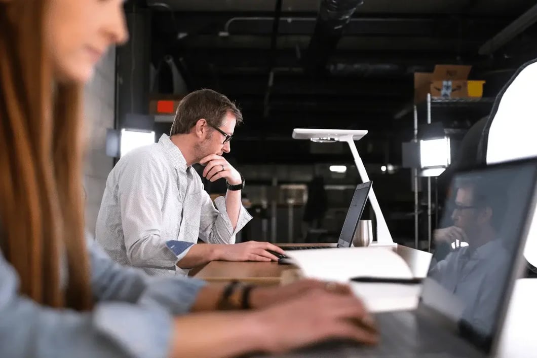 Two people focused on laptops in a dim office. The man, center, is thoughtful, chin on hand.