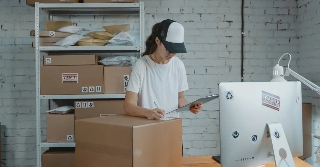 Worker scanning cardboard box in warehouse with stacked inventory, illustrating common amazon listing issues.