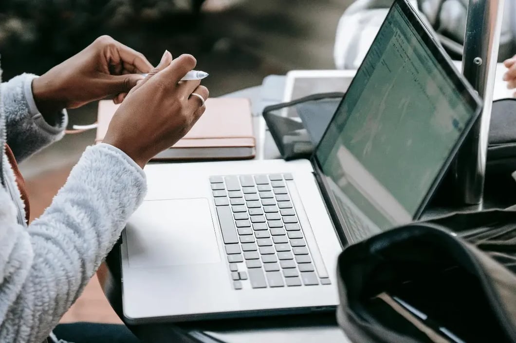 Two hands holding a pen, poised over a laptop on a desk with notebooks. 