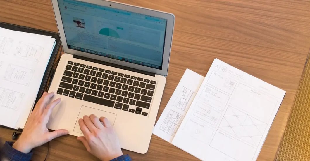 Hands typing on laptop with Amazon compliance requirements documents spread on wooden desk workspace.