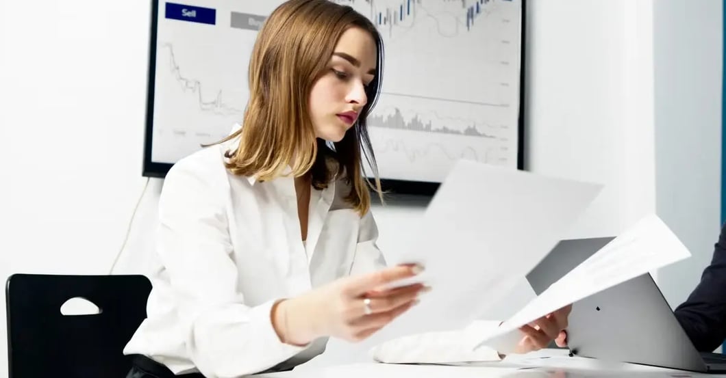 Woman reviewing financial documents at desk with stock charts on monitor, analyzing amazon holding funds.