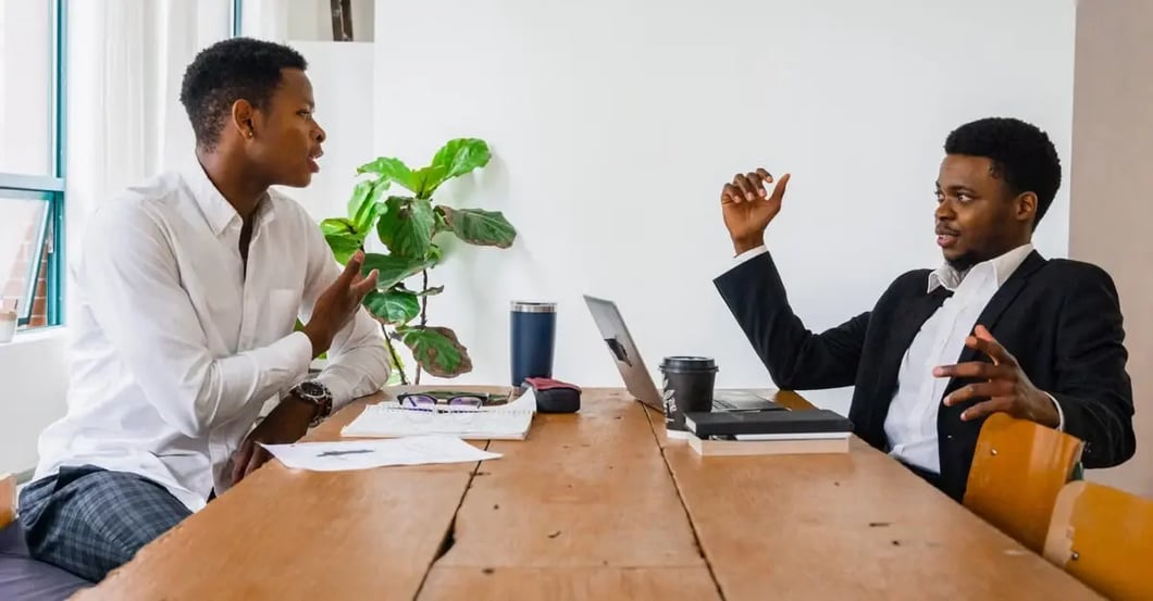 Two professionals in business attire discussing amazon consulting services at wooden table with laptop and plant.