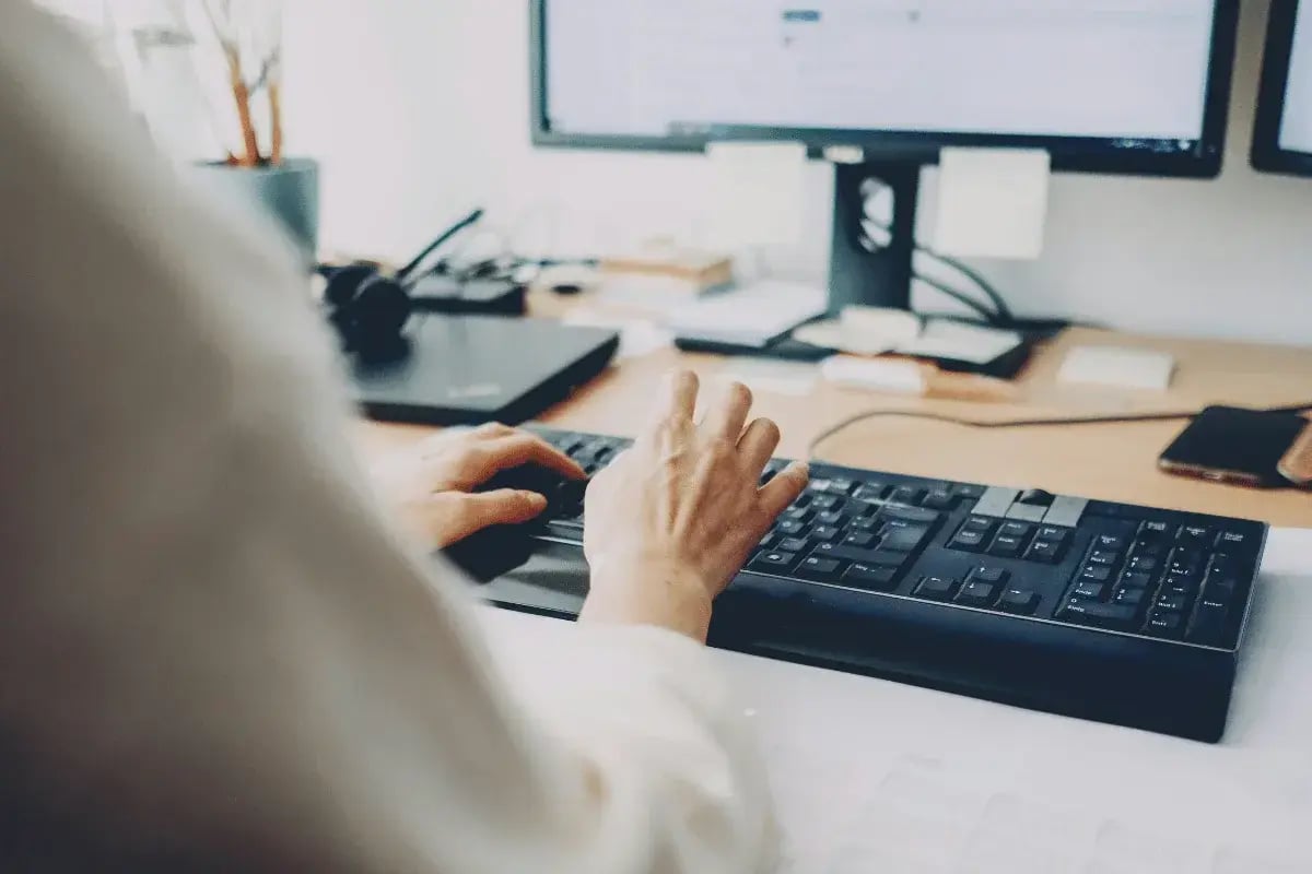 Hands typing on a black keyboard at a wooden desk, with dual monitors displaying blurred content. 