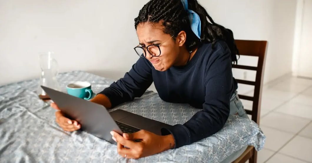 A focused woman with braided hair and glasses leans over a tablet at a kitchen table, with a turquoise mug nearby, reading intently.