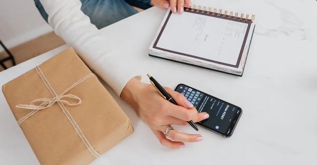 Person holding smartphone and notebook at desk with cardboard package, preparing to report listing Amazon issue.