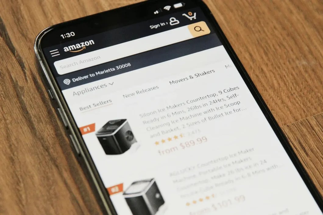 A smartphone on a wooden surface displays Amazon search results for kitchen appliances, including stainless-steel blenders with prices and ratings; a practical shopping scene.