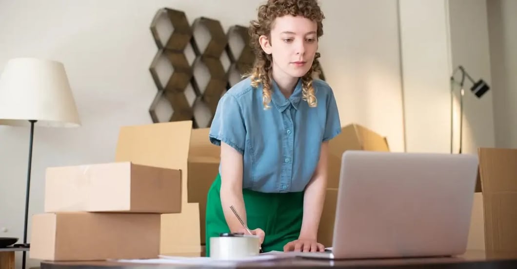 Young entrepreneur managing amazon category listing report surrounded by cardboard shipping boxes at desk with laptop.